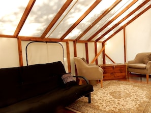 Una habitación con techo inclinado, vigas de madera, un sofá negro, un sillón color beige y una alfombra estampada.