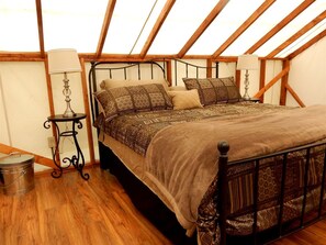Un dormitorio con techo de madera, estructura de cama de metal, ropa de cama estampada, una mesita con una lámpara y un cubo de basura.