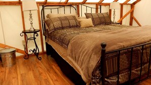 A bedroom with a wooden ceiling, a metal bed frame, patterned bedding, a small table with a lamp, and a trash can.