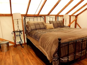 Un dormitorio con techo de madera, estructura de cama de metal, ropa de cama estampada, una mesita con una lámpara y un cubo de basura.