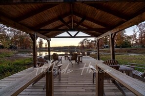 A wooden deck with Adirondack chairs and tables, overlooking a lake and autumn trees. - Exquisite Cabin with Stunning Interior, Outdoor Deck and Hot-Tub in Tennessee (Tracy City)