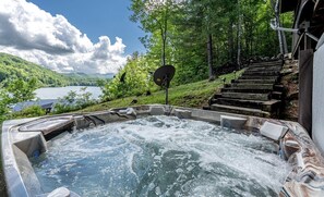 Un bain à remous avec vue sur un lac et une forêt, entouré de marches en pierre et d’arbres.