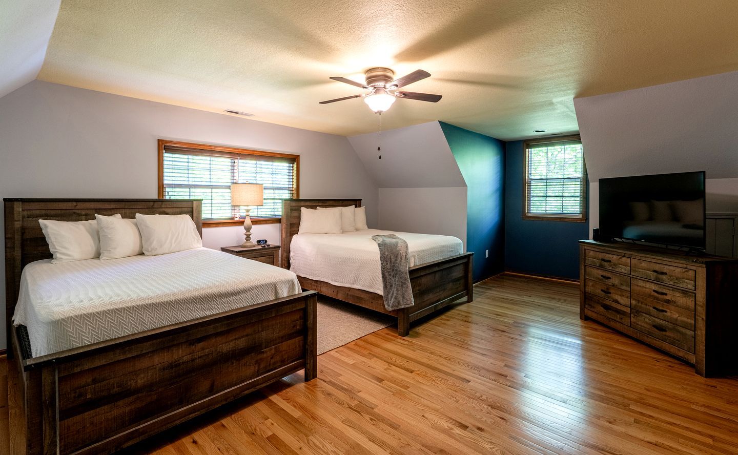 A bedroom with two beds, a ceiling fan, a dresser, a television, and windows with blinds.