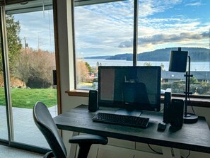 A computer desk with a monitor, keyboard, and speakers, positioned by a window with a view of a lake and hills. - Peaceful Cabin with Bay Views and Outdoor Hot-Tub in Washington (Anacortes)