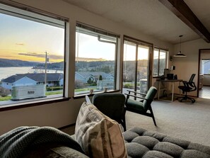 A living room with a view of a river and houses, a green armchair, a sofa, and a desk with a computer. - Peaceful Cabin with Bay Views and Outdoor Hot-Tub in Washington (Anacortes)