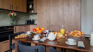 Une table de cuisine moderne, en bois, est dressée pour le petit-déjeuner : on y trouve un panier de fruits, des viennoiseries et une théière.