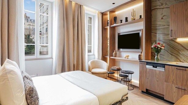 Une chambre d’hôtel comprenant un lit, un coin salon avec un fauteuil et de petites tables, une cuisine intégrée avec un téléviseur, et une vue sur la ville grâce à de grandes fenêtres.