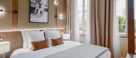 Camera d'albergo con un letto grande, una testiera con luci a parete, un quadro incorniciato sopra il letto e una finestra con tende.
