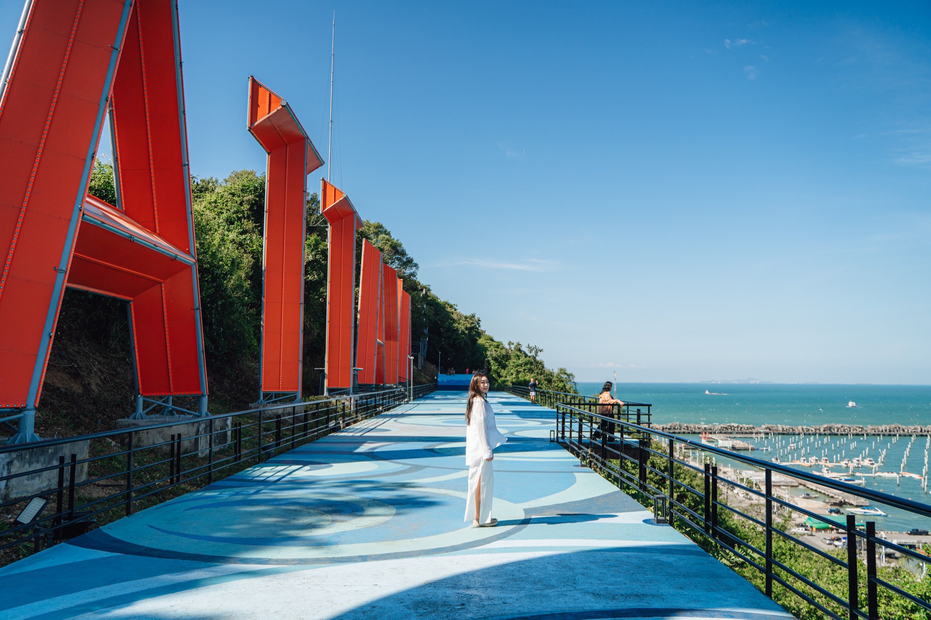 A bridge with red structures, a person standing on the walkway, and a view of the sea.