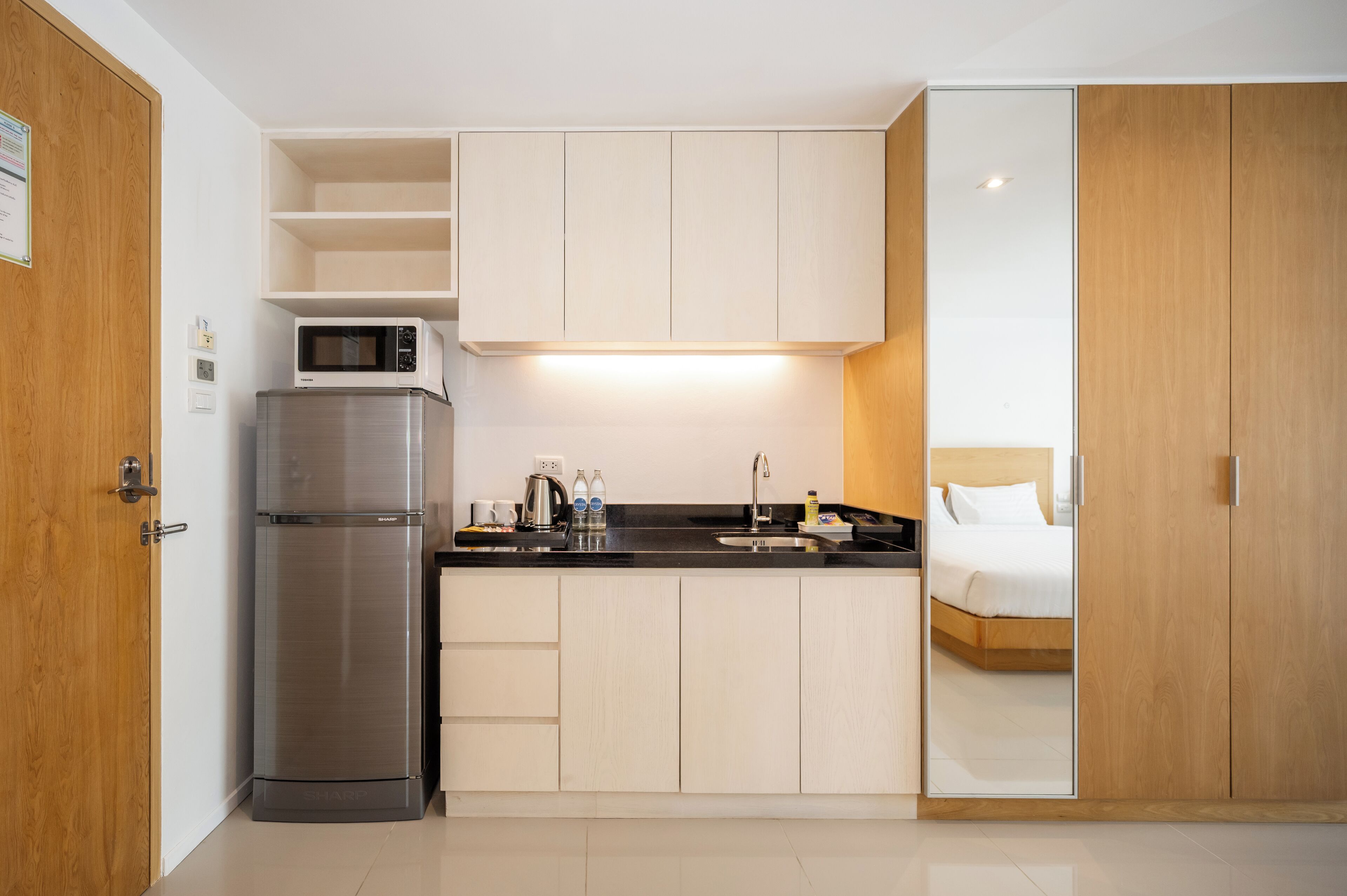 A compact kitchen with a refrigerator, microwave, and sink, adjacent to a bedroom with a bed and wooden wardrobe.