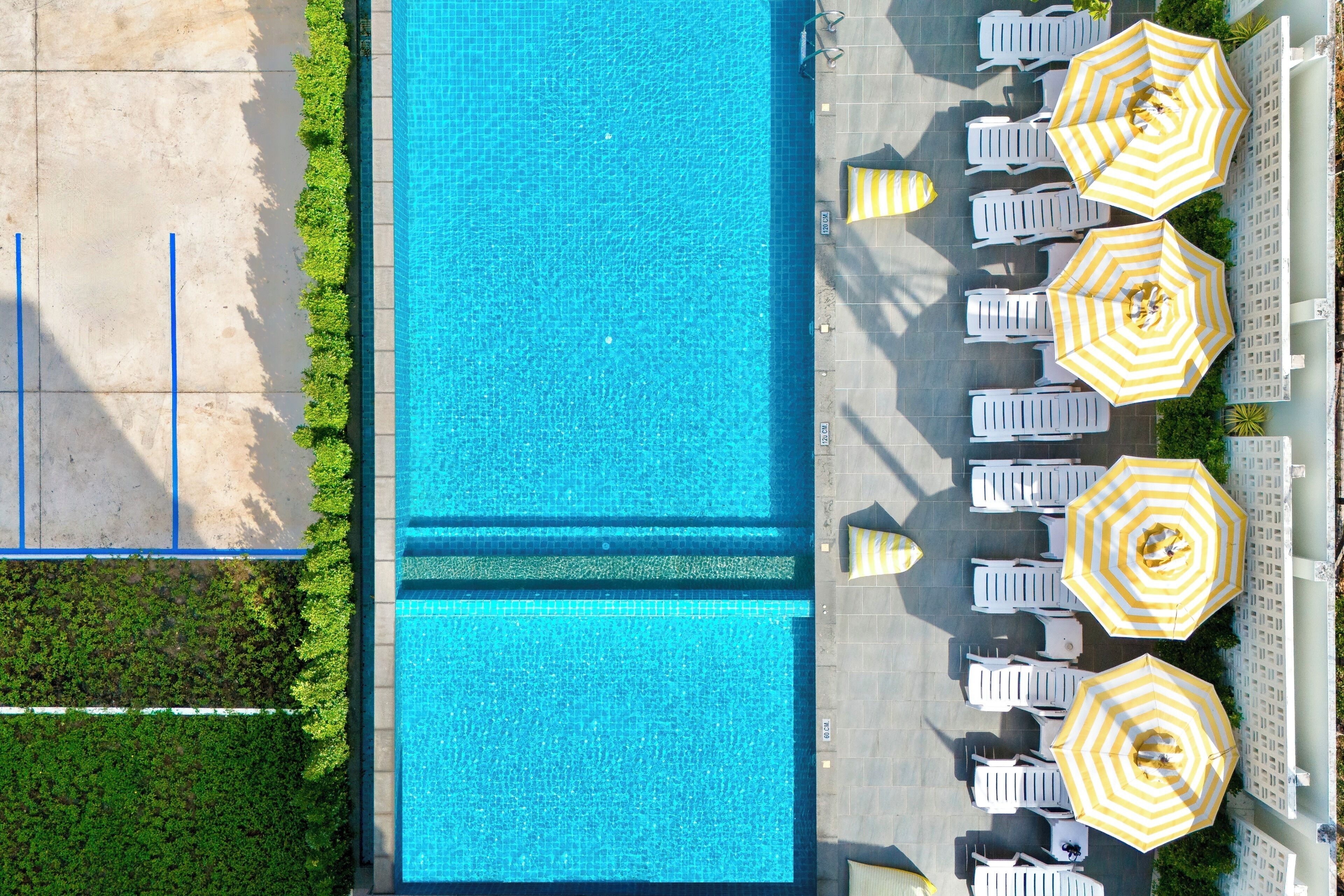 An aerial view of a swimming pool with adjacent lounge chairs and umbrellas, surrounded by greenery and a tiled walkway.