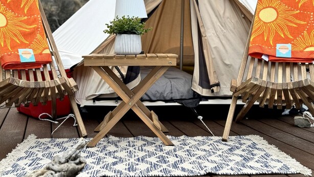 Een tent met een tafel en stoelen, een klein plantje en een vloerkleed op een houten terras.