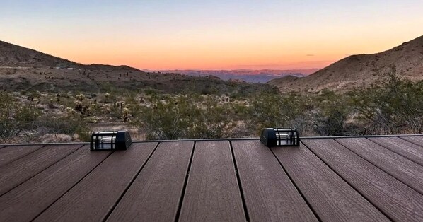 Una terraza de madera con dos objetos negros, probablemente altavoces, con vistas a un paisaje montañoso de vegetación escasa.