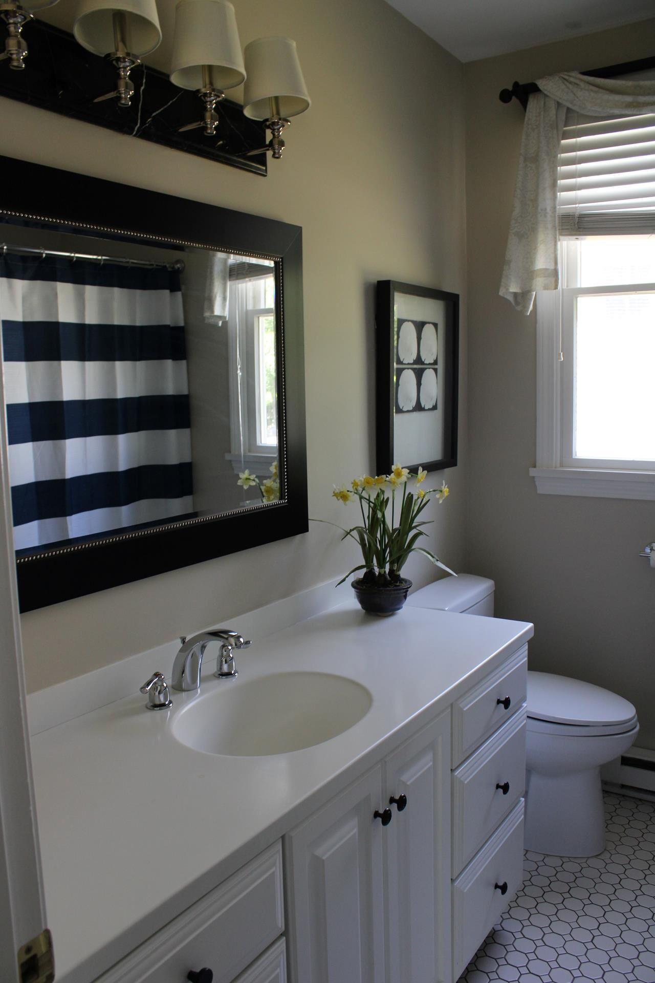 Une salle de bain avec un meuble-lavabo blanc, un grand miroir, un rideau de douche à rayures noires et blanches, un pot de jonquilles et des toilettes.