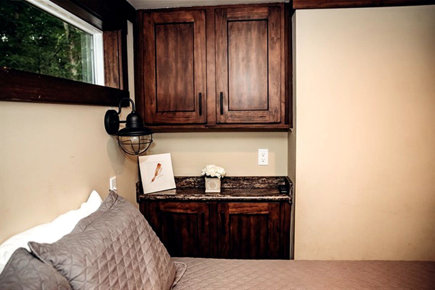 A bedroom with a bed, a nightstand, a wooden cabinet above the nightstand, and a window with a view of trees.