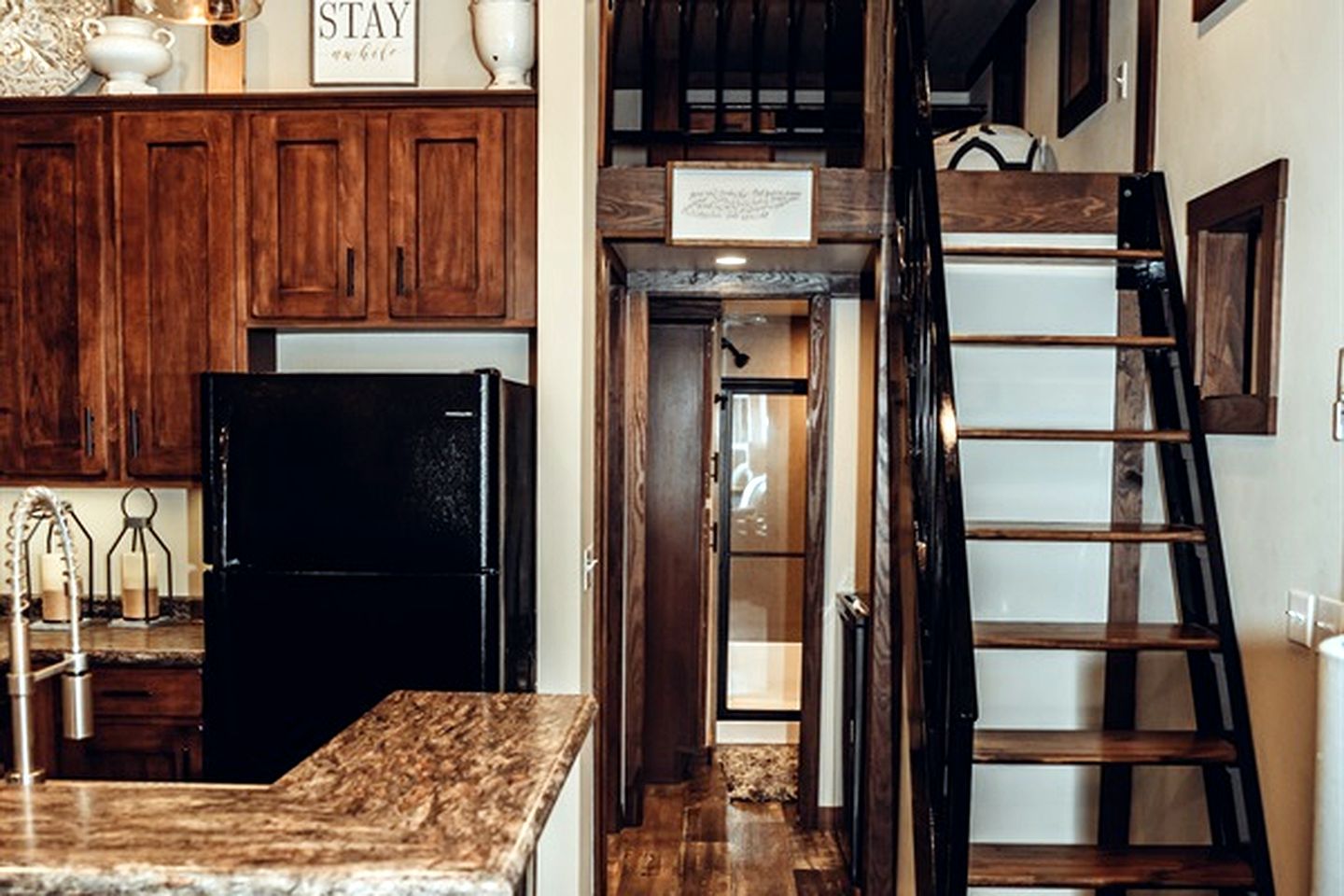A kitchen with wooden cabinets, a black refrigerator, a marble countertop, a ladder, and a sign above the door.