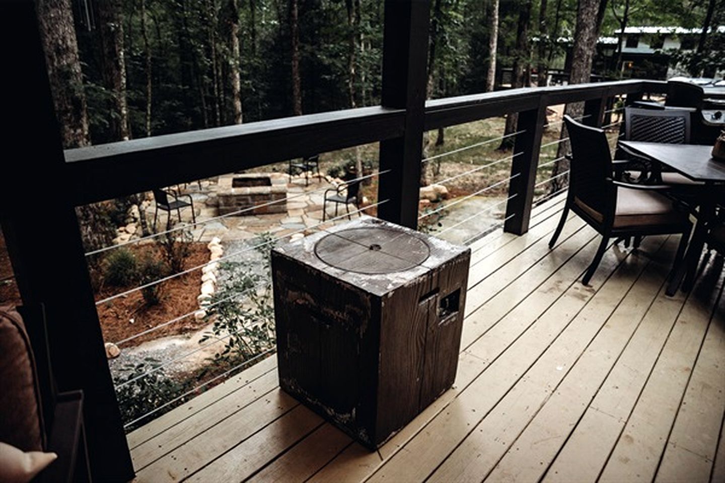 A wooden deck with a stone fire pit, surrounded by chairs and tables, overlooking a forested area.