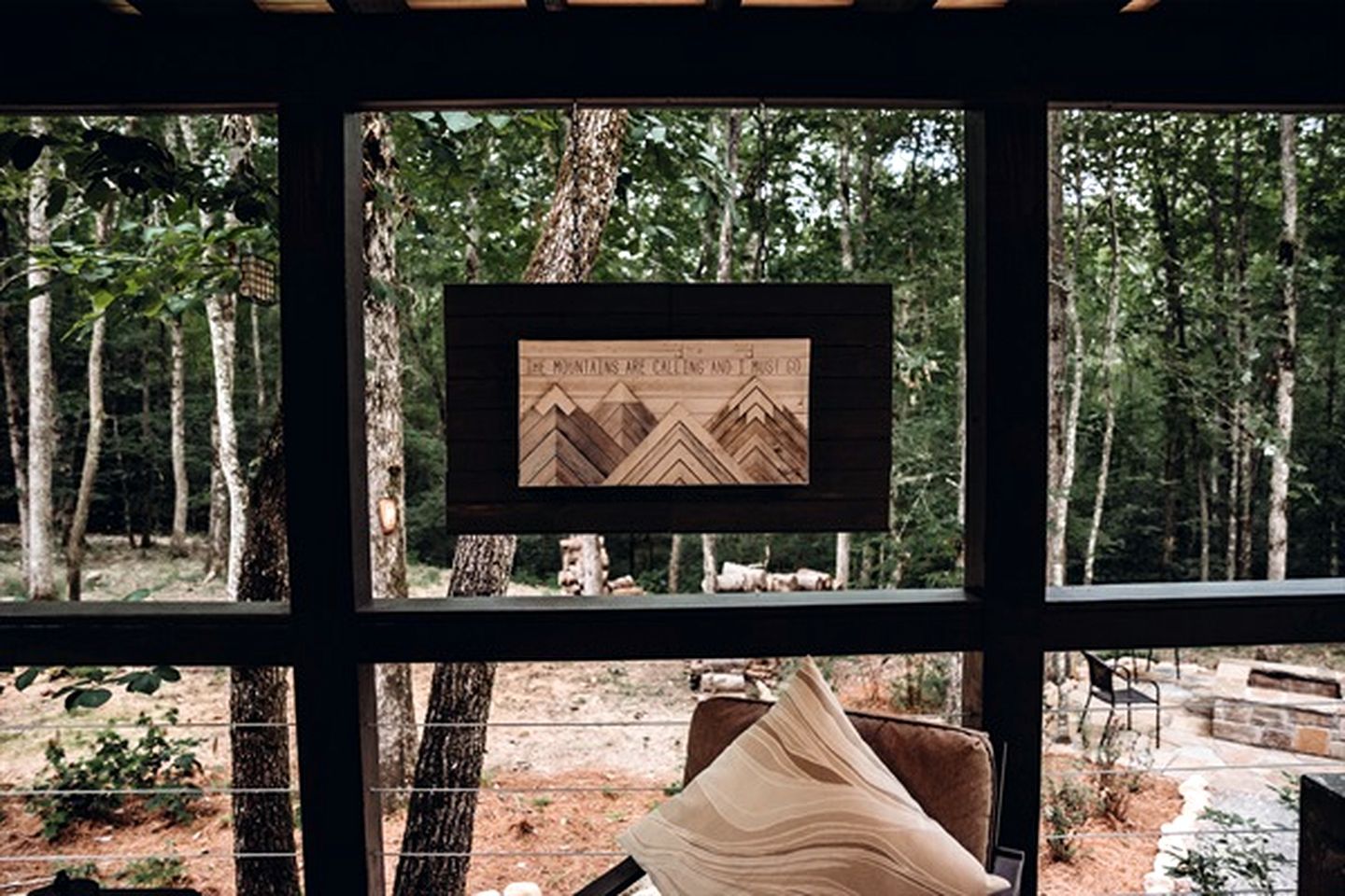 Una obra de arte enmarcada que muestra montañas y el texto 'THE MOUNTAINS ARE CALLING AND I HEAR THEM' en una habitación con vista a un bosque.
