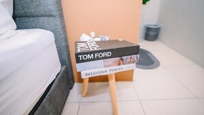 A small table with a book titled 'Tom Ford' and 'Delicious Places' on it, placed in a room with a grey sofa and a white bed. - Swing & Pillows at Penang Road (George Town)