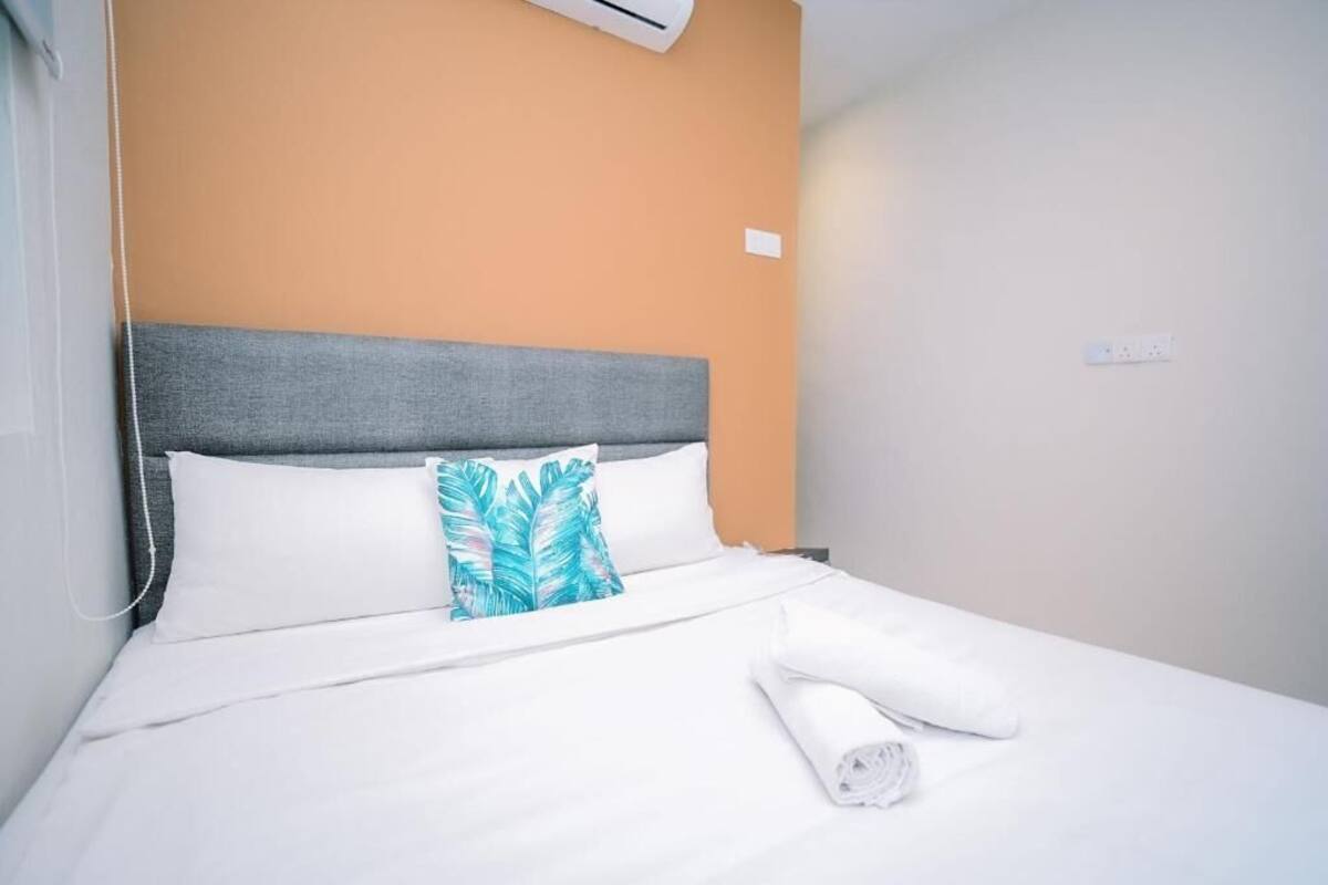 Habitación de hotel con cama, ropa de cama blanca, cabecero gris y una sola almohada con diseño de hojas azules.