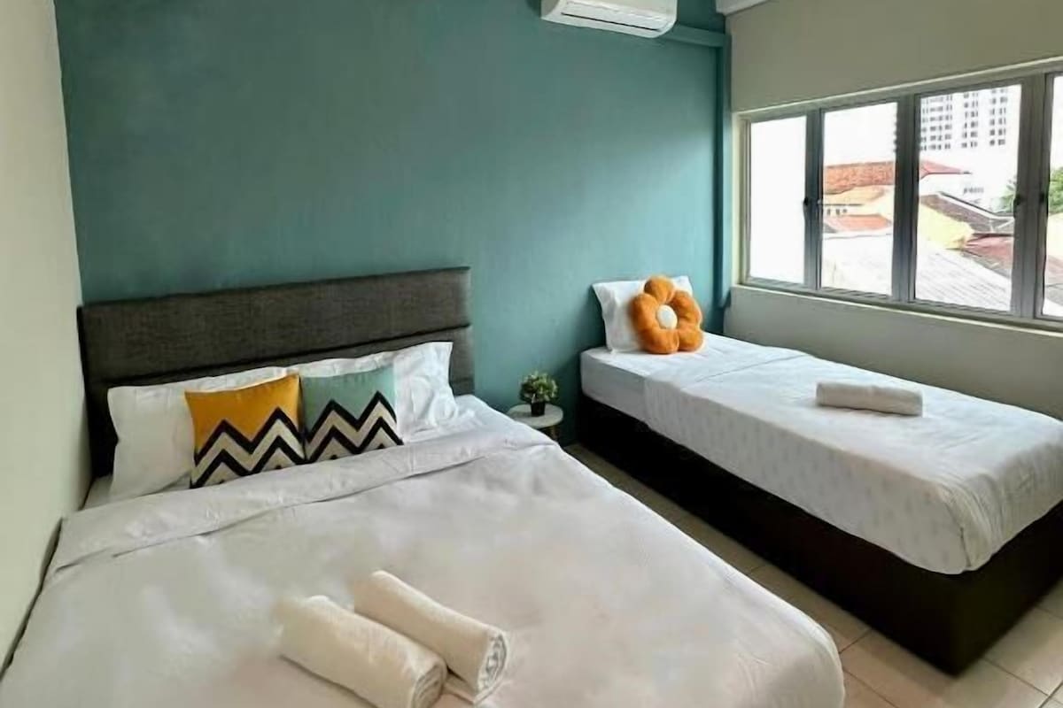 Habitación de hotel con dos camas, paredes color turquesa, un equipo de aire acondicionado y ventana con vista a edificios.