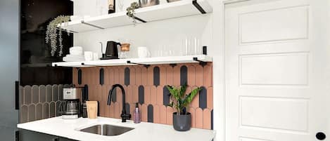 Une cuisine moderne avec un plan de travail blanc, des meubles noirs, une machine à café, un évier et une plante en pot.