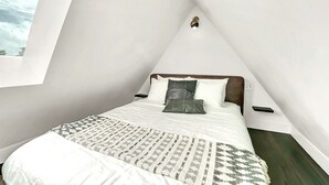 Ein Schlafzimmer mit schrägen Wänden, einem Bett mit weißer Bettwäsche, einer gemusterten Tagesdecke und einem Nachttisch mit Telefon.