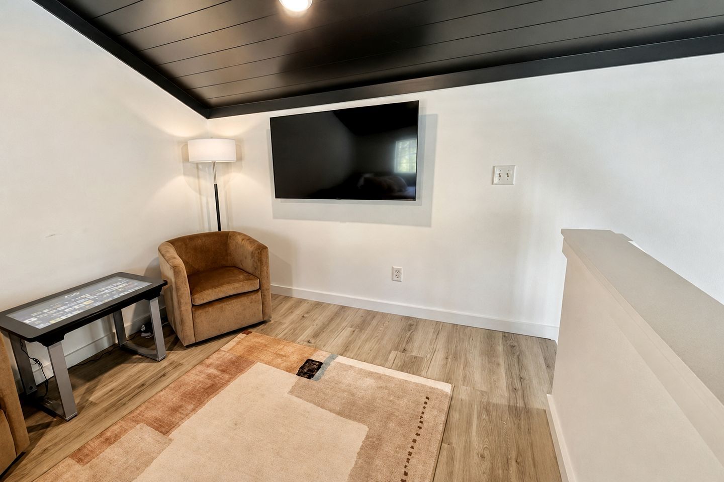 A room with a flat-screen TV mounted on the wall, a brown armchair, a small table with a digital interface, and a patterned rug on the wooden floor.