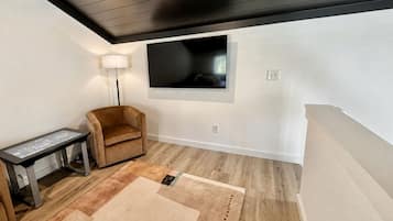 A room with a flat-screen TV mounted on the wall, a brown armchair, a small table with a digital interface, and a patterned rug on the wooden floor.