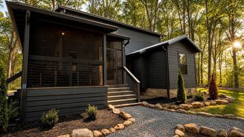 A modern two-story cabin with a covered porch, a stone pathway, and a forested backdrop.