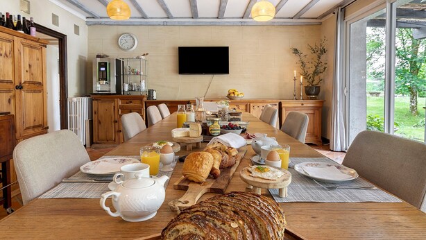 Une table en bois, dans une pièce équipée d’un téléviseur et d’armoires en bois, est dressée pour le petit-déjeuner avec du pain, des œufs et diverses boissons.