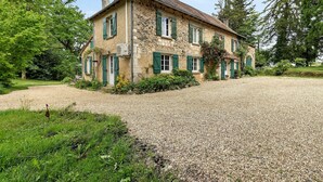 Une maison en pierre avec des volets verts, une allée en gravier et une végétation luxuriante tout autour.