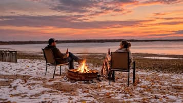 Two people sitting by a campfire on a snow-covered ground with a view of a body of water and a sunset.