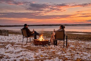 Two people sitting by a campfire on a snow-covered ground with a view of a body of water and a sunset.