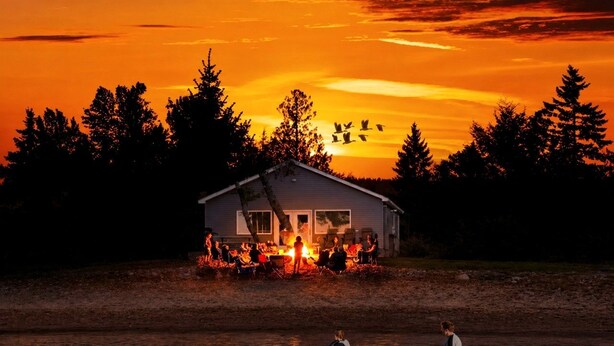A cabin by a lake at sunset with people gathered outside.