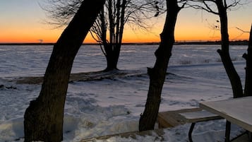 A wooden deck with a table, surrounded by bare trees, overlooking a frozen body of water during sunset.