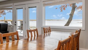 A dining area with a large table and chairs, a view of the beach, and a tree with autumn leaves.