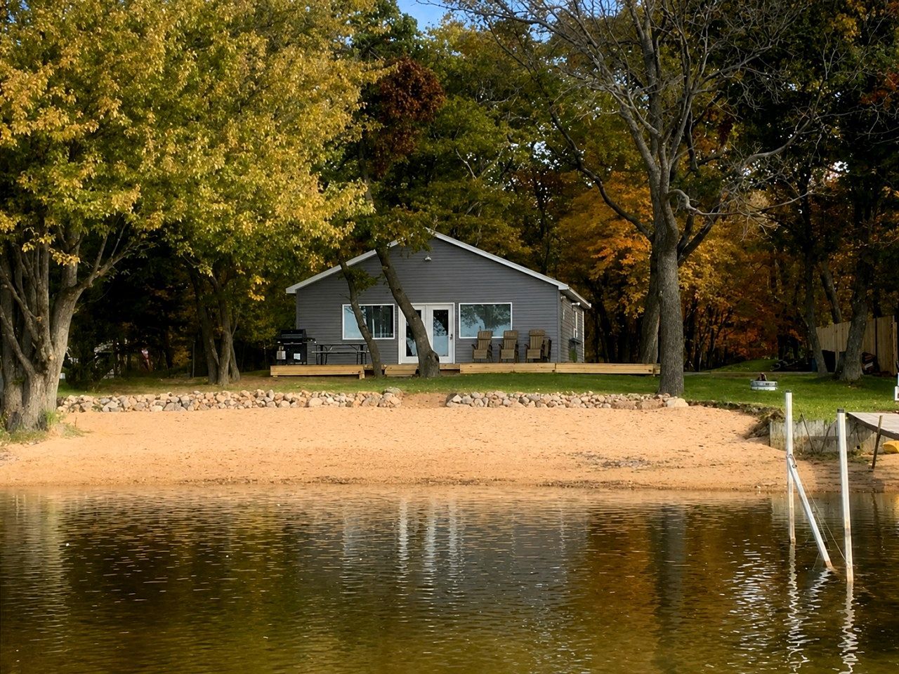 A lakeside cabin with a dock, surrounded by trees and a sandy beach.