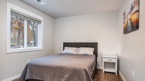 A bedroom with a bed, a nightstand, a window with blinds, and a painting on the wall. - Waterfront Cabin with BBQ, Picnic Space and Amazing Sunsets in Michigan (Au Gres)