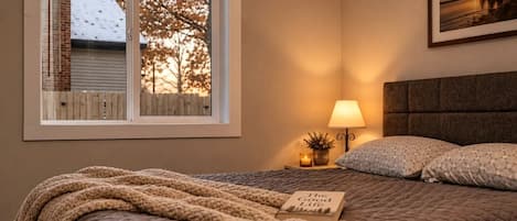 Un dormitorio con cama, lámpara de noche, un cuadro en la pared y ventana con vistas a árboles y un edificio.