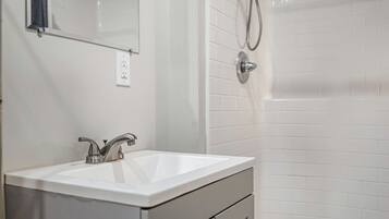 A bathroom with a white sink, a mirror, a shower with a handheld showerhead, and a tiled wall.