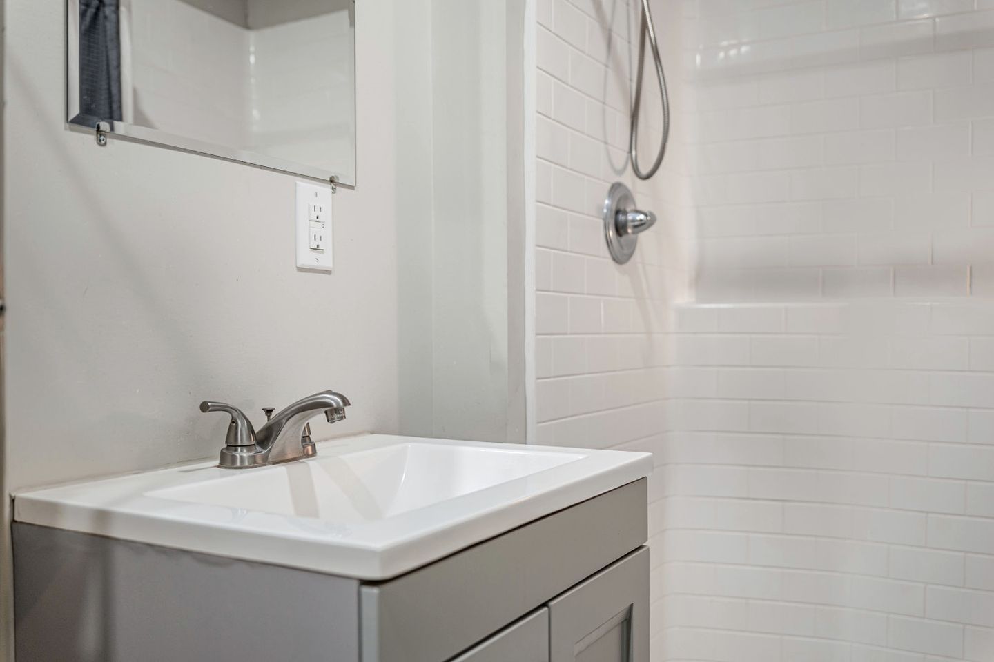 A bathroom with a white sink, a mirror, a shower with a handheld showerhead, and a tiled wall.