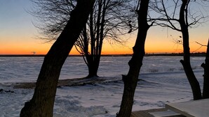 A wooden deck with a table, surrounded by bare trees, overlooking a frozen body of water during sunset.