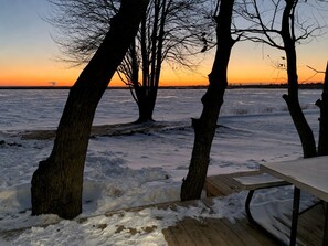 A wooden deck with a table, surrounded by bare trees, overlooking a frozen body of water during sunset.