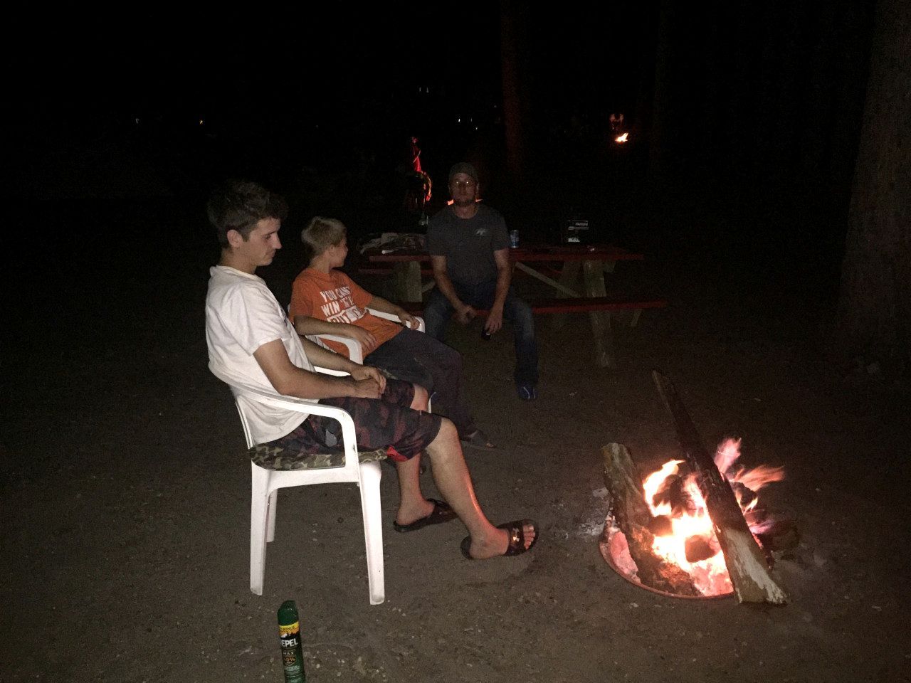 Trois personnes sont assises à l’extérieur, autour d’un feu de camp, pendant la nuit.