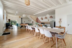 A spacious dining area with a long wooden table and white chairs, a staircase leading to another level, and a kitchen in the background.