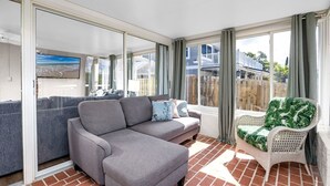 Eine sonnendurchflutete Terrasse mit einer grauen Eckcouch, einem Korbstuhl mit Kissen mit tropischem Muster und einem großen Fenster mit Vorhängen.