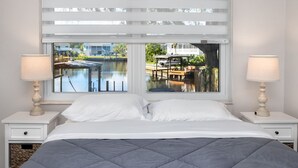 Ein Schlafzimmer mit Bett, zwei Nachttischen, einem Fenster mit Jalousien und Blick auf ein Gewässer und Gebäude.