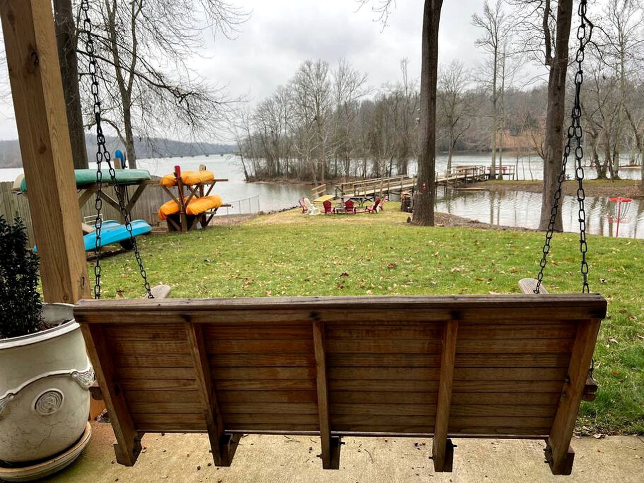 A wooden swing chair hanging from a wooden structure, a small table with chairs, and a view of a lake with trees in the background.