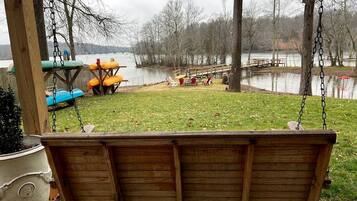 A wooden swing chair hanging from a wooden structure, a small table with chairs, and a view of a lake with trees in the background.